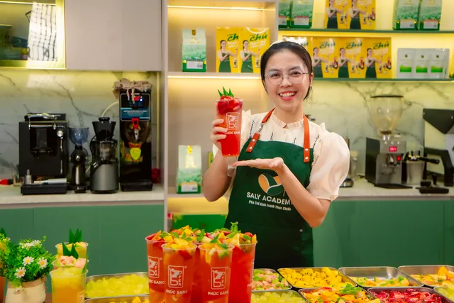 Cô Ly Phạm mặc tạp dề cười, cầm ly trà trái cây tại quầy pha chế có máy pha cà phê