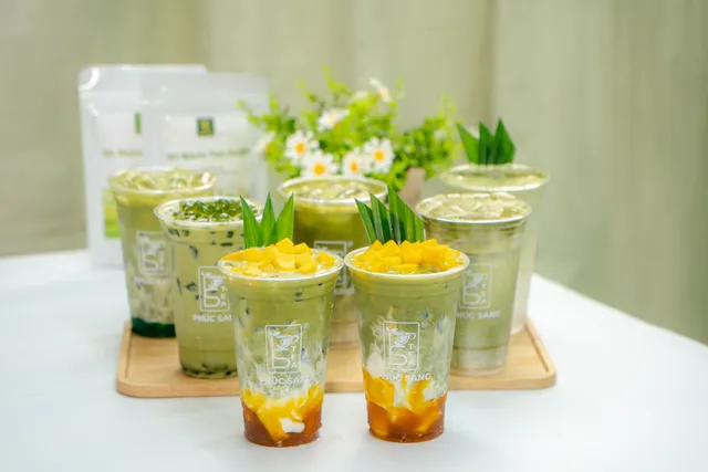 Nhiều ly matcha latte và matcha xoài xếp trên khay gỗ cạnh bình hoa